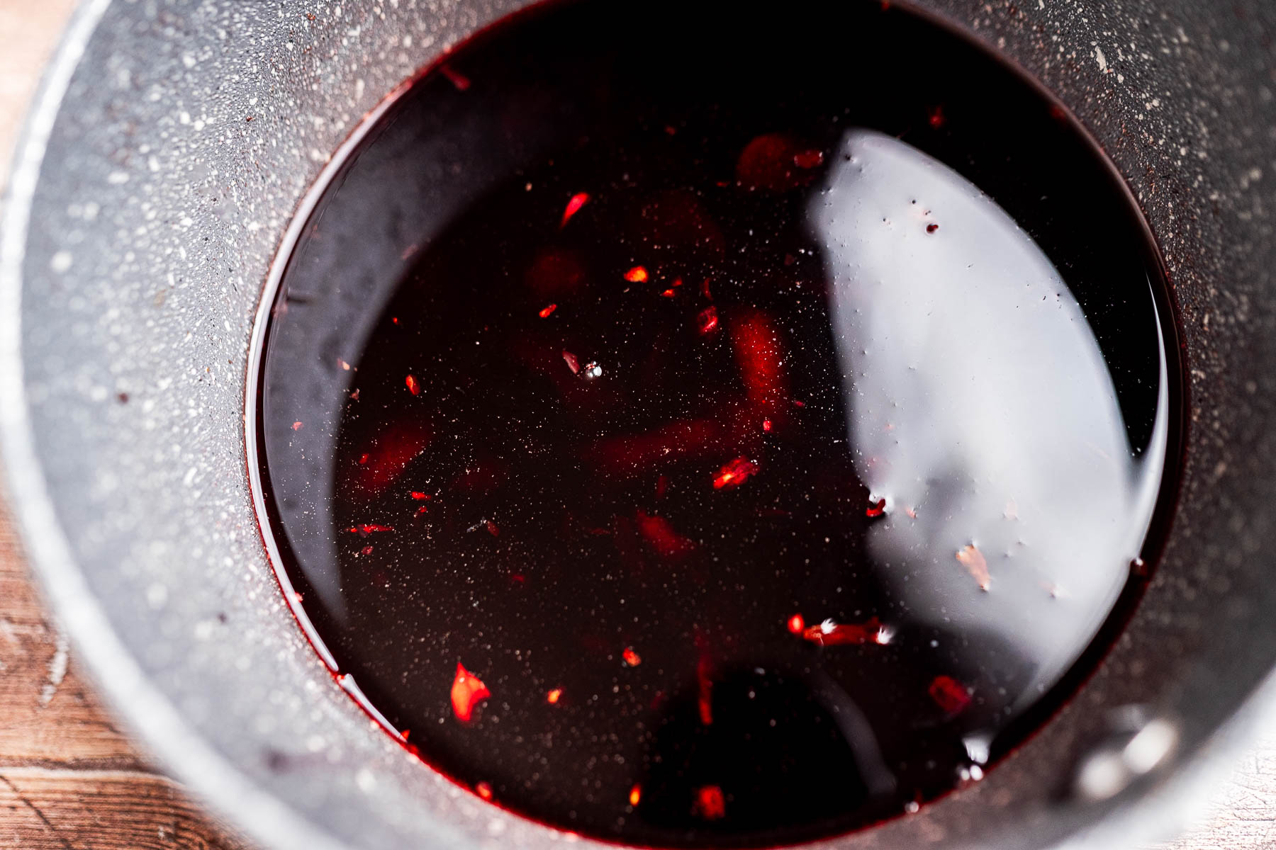 A close-up of hibiscus syrup, its dark red liquid and small red fragments nestled in a speckled gray metal bowl on a wooden surface.