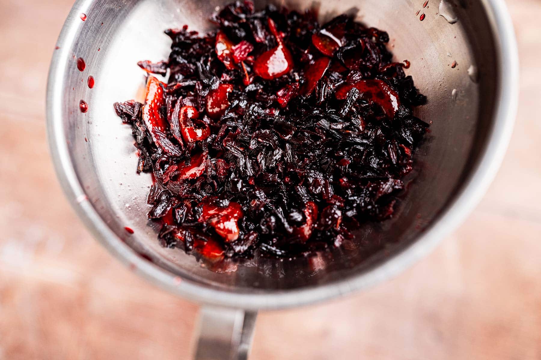 A close-up of a metal bowl containing a mixture of cooked black rice and red bell pepper pieces, lightly drizzled with hibiscus syrup.