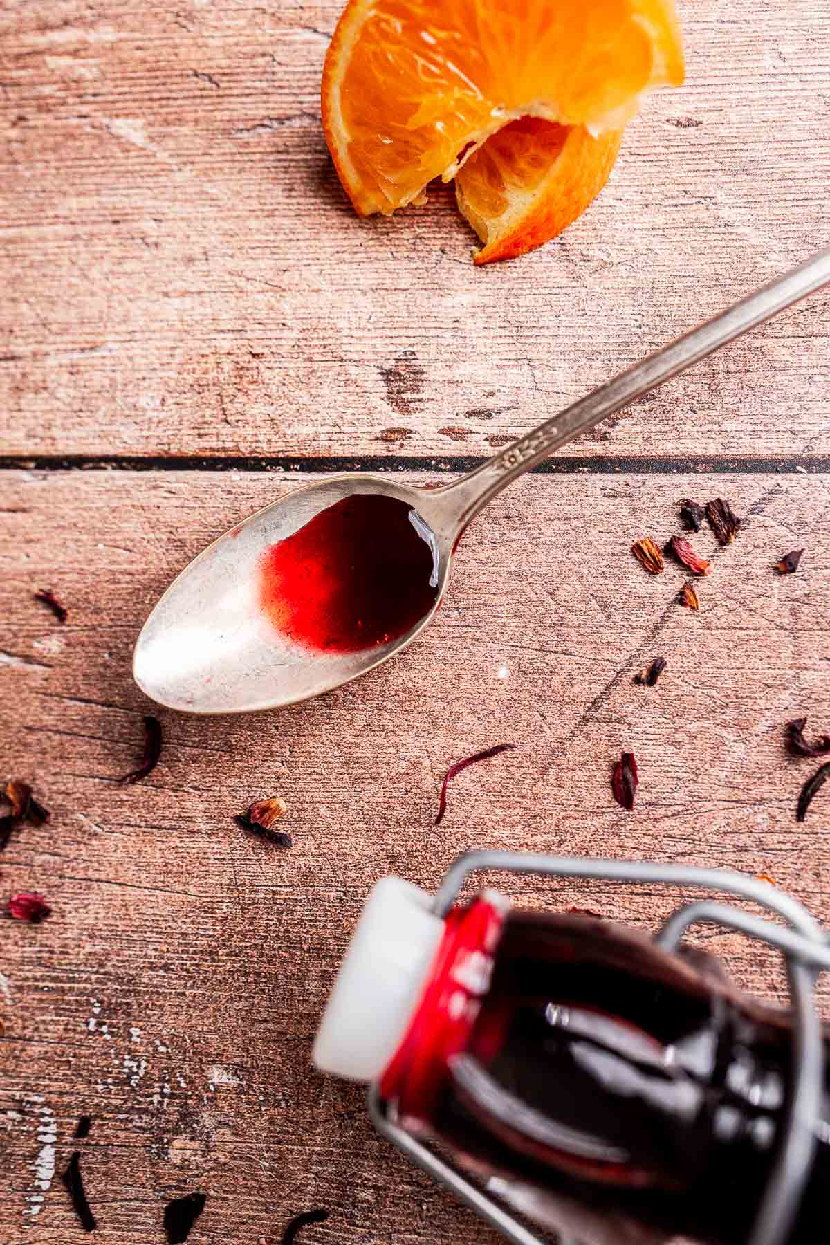 A spoon with hibiscus syrup, a small glass bottle filled with red liquid, dried flower petals, and orange slices on a wooden surface.