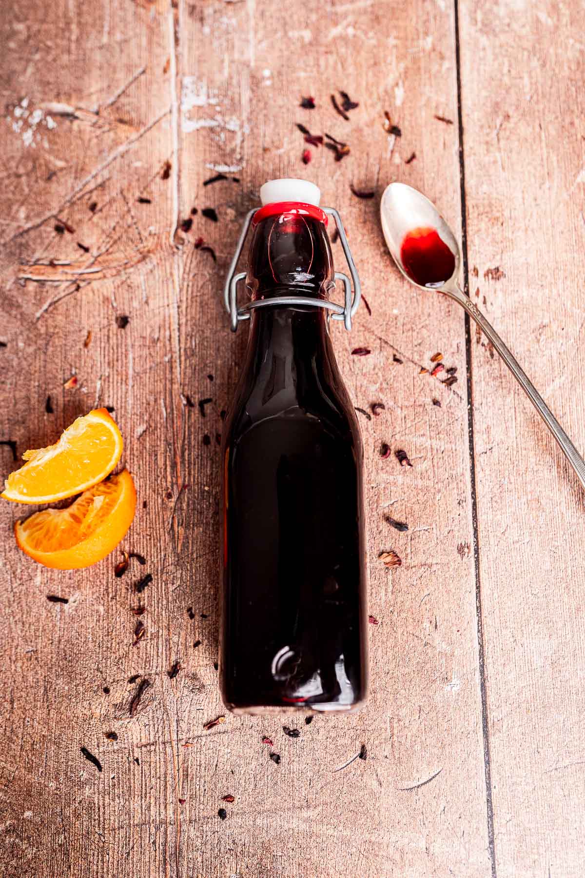 A glass bottle filled with dark hibiscus syrup sits on a wooden surface, next to orange slices, scattered dried flowers, and a spoon with some of the vibrant liquid.