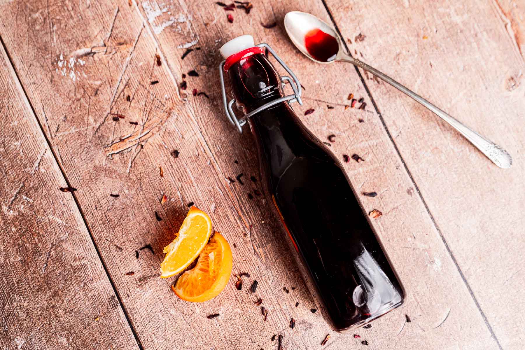 A glass bottle filled with dark hibiscus syrup, a spoon with red residue, two orange slices, and scattered dried petals on a wooden surface.