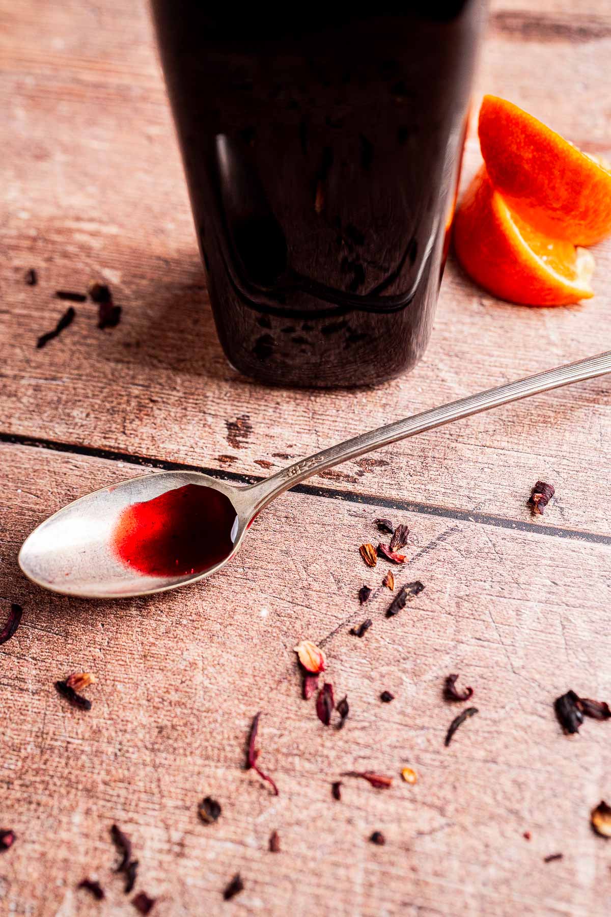A spoon with dark red hibiscus syrup sits on a wooden surface near scattered dried herbs, a glass of dark beverage, and orange slices.