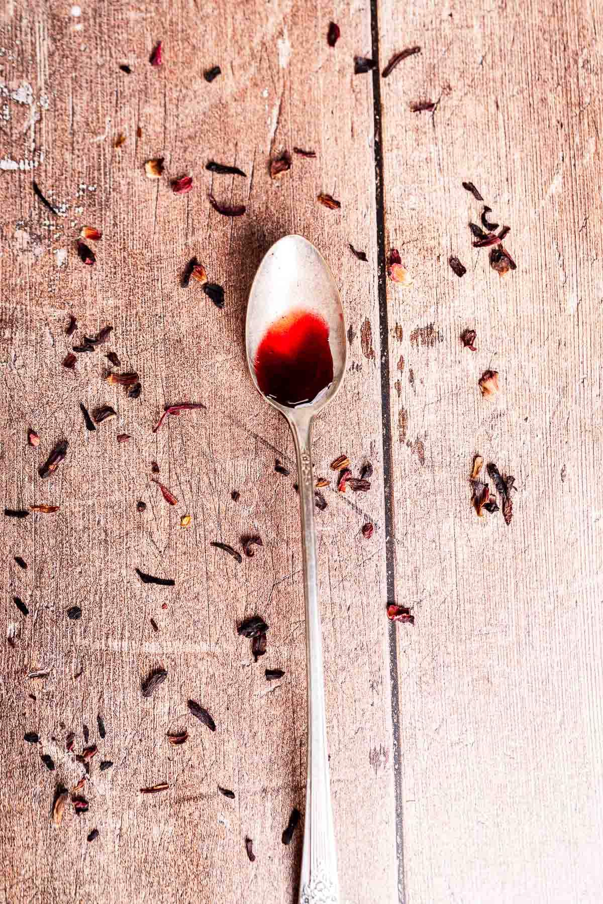 A silver spoon with a small amount of hibiscus syrup rests on a wooden surface scattered with dried hibiscus petals.