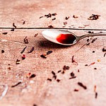 A silver spoon with a small amount of hibiscus syrup rests on a wooden surface scattered with dried tea leaves.
