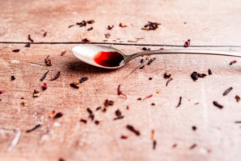 A silver spoon with a small amount of hibiscus syrup rests on a wooden surface scattered with dried tea leaves.