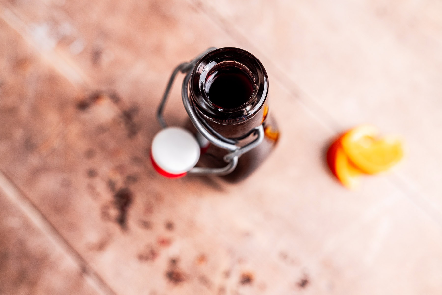 Top view of an open brown glass bottle with a swing-top cap, filled with hibiscus syrup, placed on a wooden surface next to a sliced orange.
