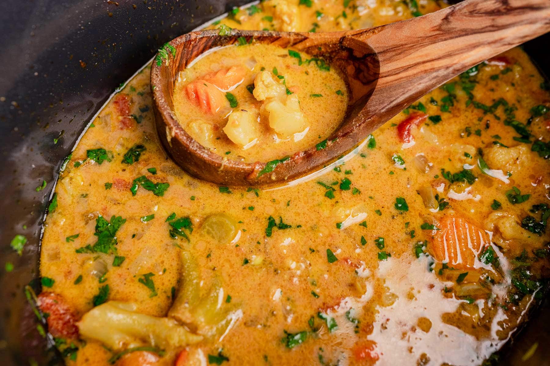 A wooden spoon lifts slow cooker vegetable curry with carrots, cauliflower, and herbs from a pot filled with creamy, orange-colored broth.