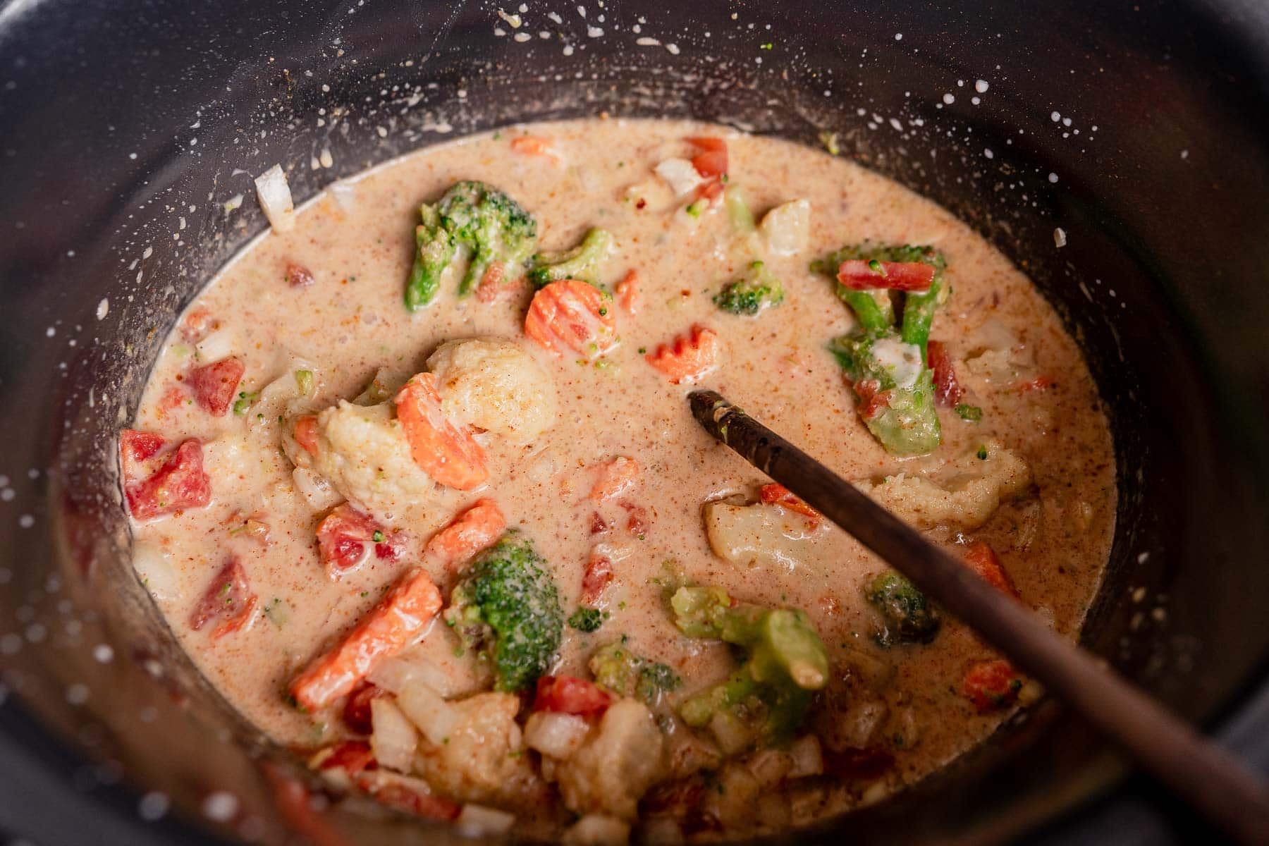 A close-up of a creamy slow cooker vegetable curry with broccoli, carrots, cauliflower, and red bell pepper being stirred in a pot.