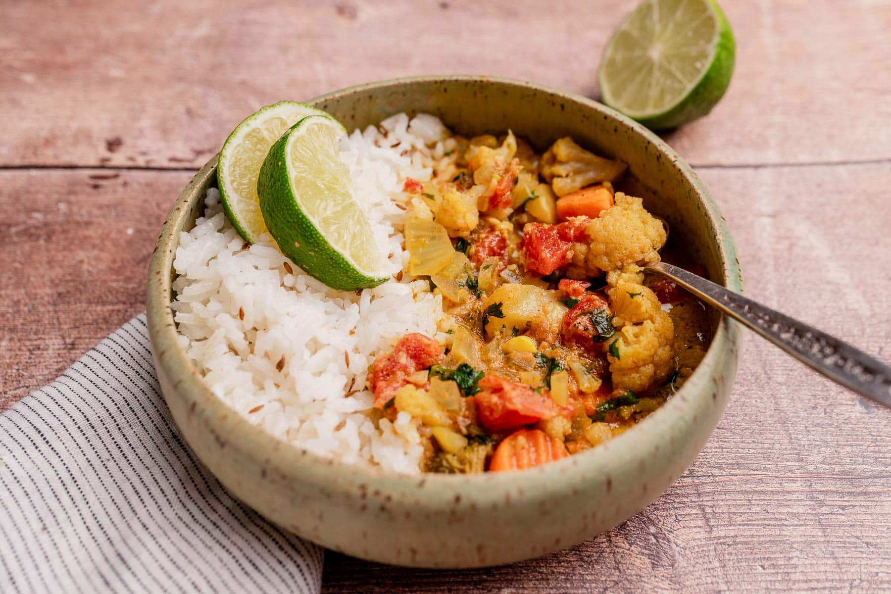 A bowl of white rice and slow cooker vegetable curry garnished with lime slices, set on a striped napkin with a halved lime in the background.