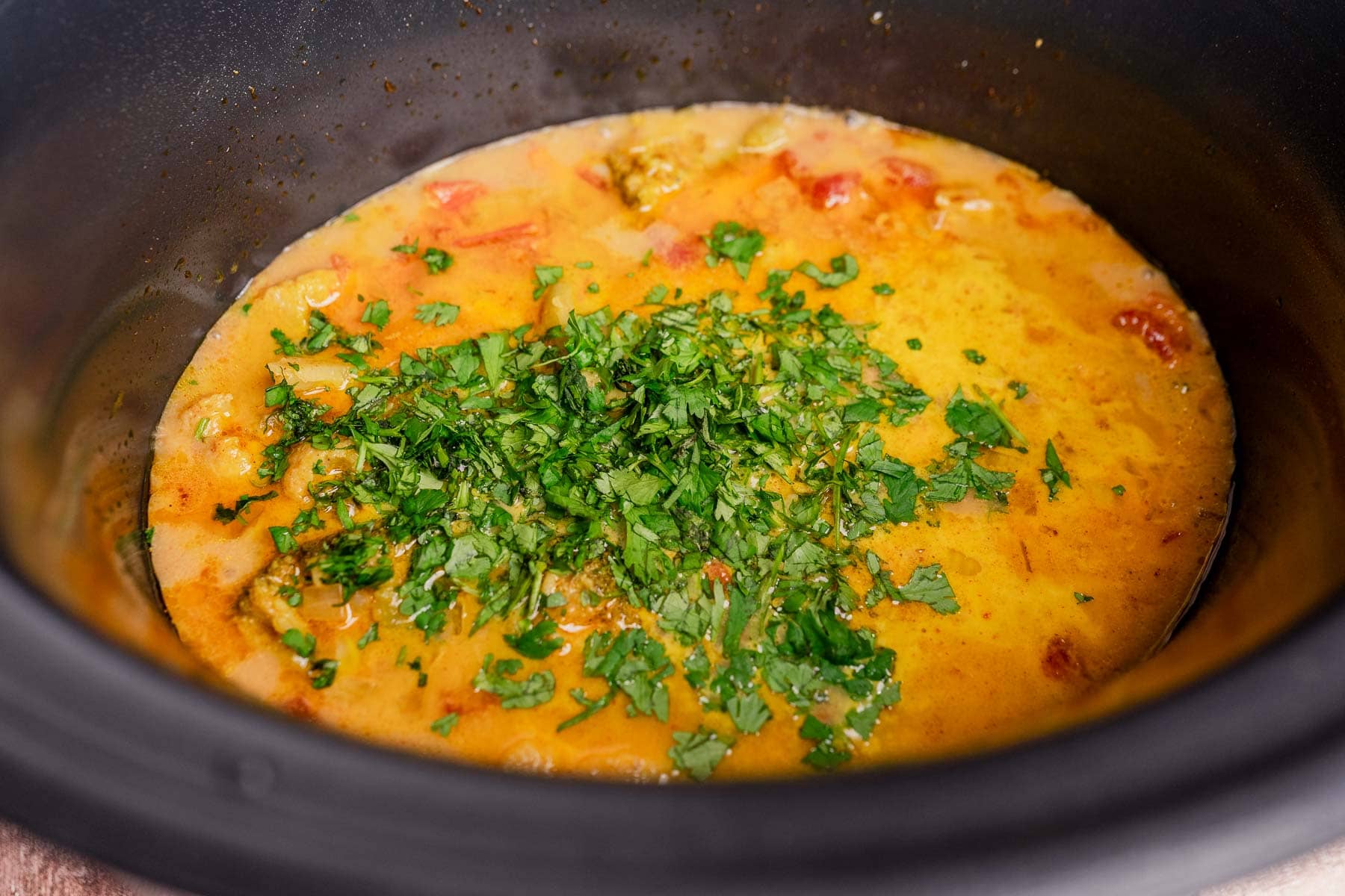 A yellow and orange slow cooker vegetable curry with chopped tomatoes and fresh cilantro sits in a black slow cooker.