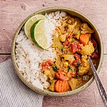 A bowl of white rice and slow cooker vegetable curry garnished with lime slices sits on a wooden surface beside a striped cloth napkin.