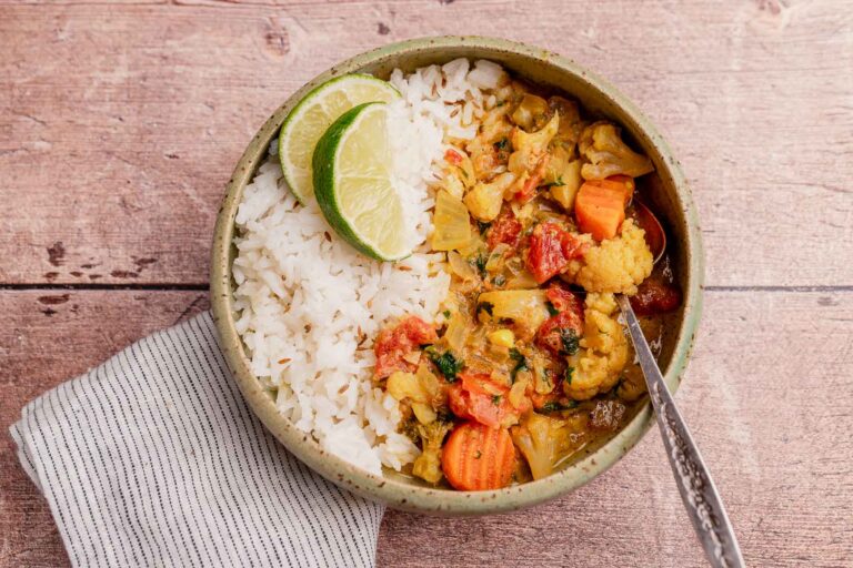 A bowl of white rice and slow cooker vegetable curry garnished with lime slices sits on a wooden surface beside a striped cloth napkin.