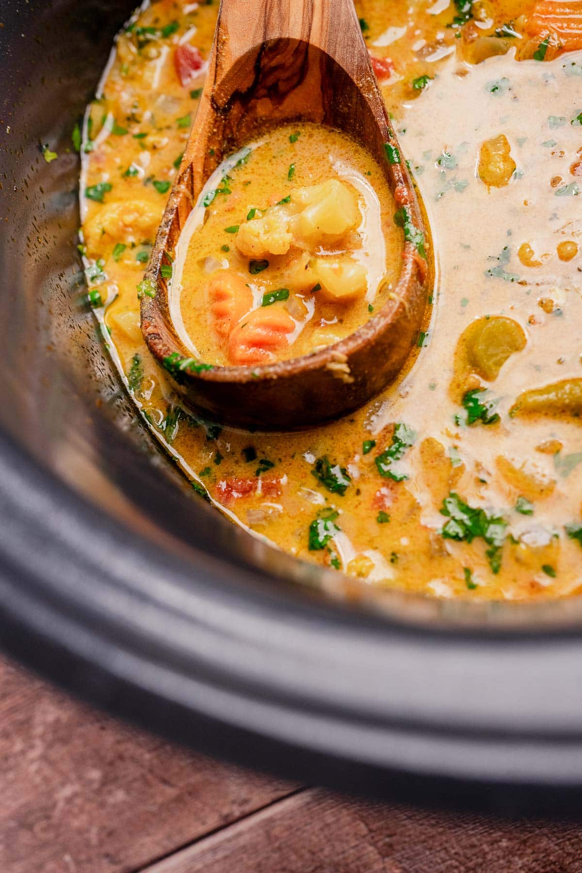 A wooden spoon rests in a pot of creamy slow cooker vegetable curry soup with visible vegetables and herbs.