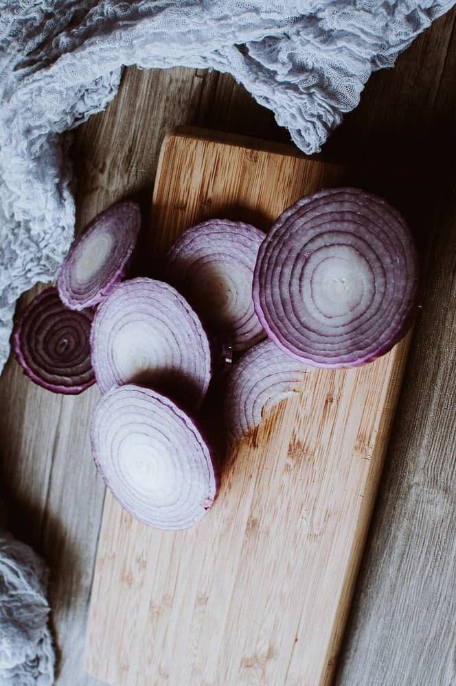Several slices of red onion are arranged on a wooden cutting board, with a textured cloth partially visible in the background.