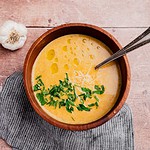 A wooden bowl of creamy garlic soup garnished with chopped herbs, grated cheese, and olive oil, with a spoon inside. A garlic bulb and napkin rest beside the bowl on a wooden surface.