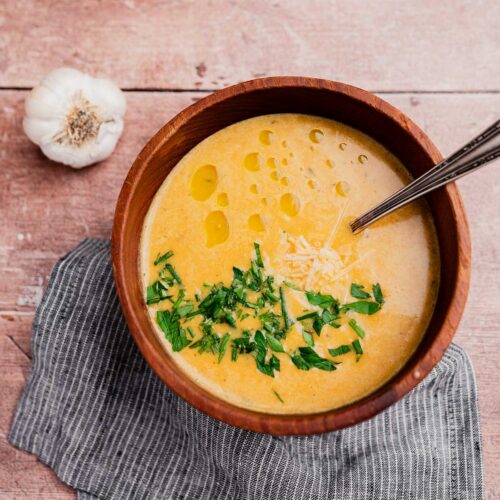 A wooden bowl of creamy garlic soup garnished with chopped herbs, grated cheese, and olive oil, with a spoon inside. A garlic bulb and napkin rest beside the bowl on a wooden surface.