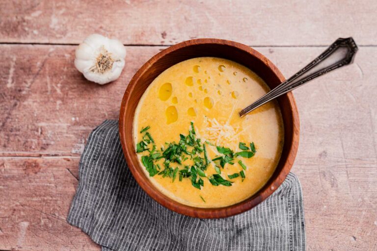 A wooden bowl of creamy garlic soup garnished with chopped herbs, grated cheese, and olive oil, with a spoon inside. A garlic bulb and napkin rest beside the bowl on a wooden surface.