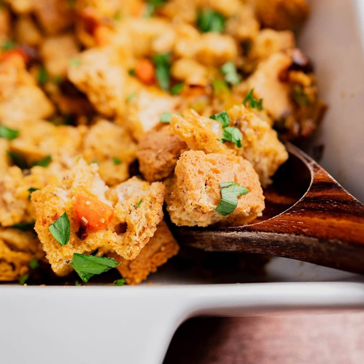 Close-up of a wooden spoon scooping gluten free cornbread stuffing with chopped herbs and vegetables from a white dish.