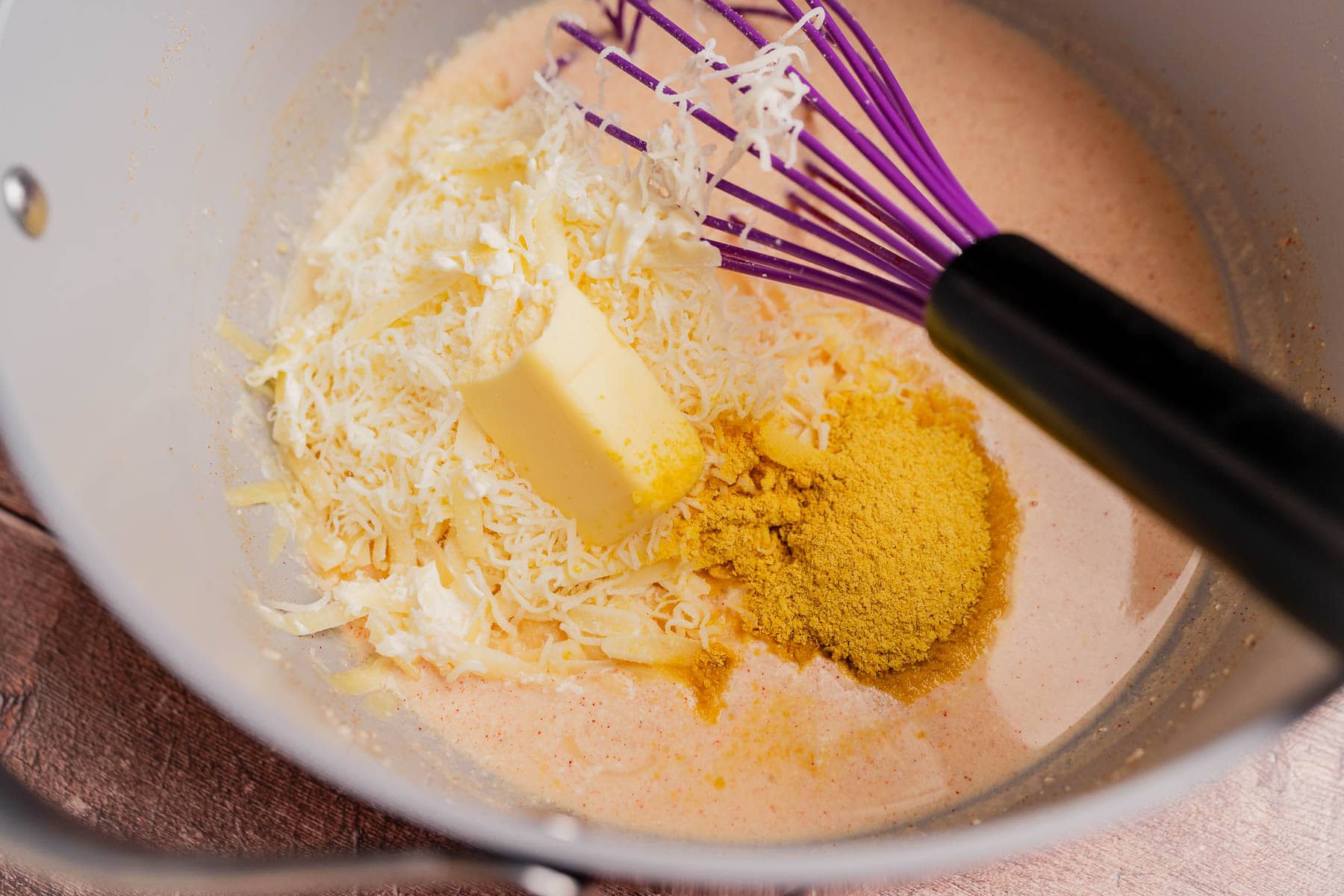 A saucepan containing shredded cheese, a block of butter, powdered seasoning, and cream is being mixed with a purple whisk for creamy gluten free mac and cheese.