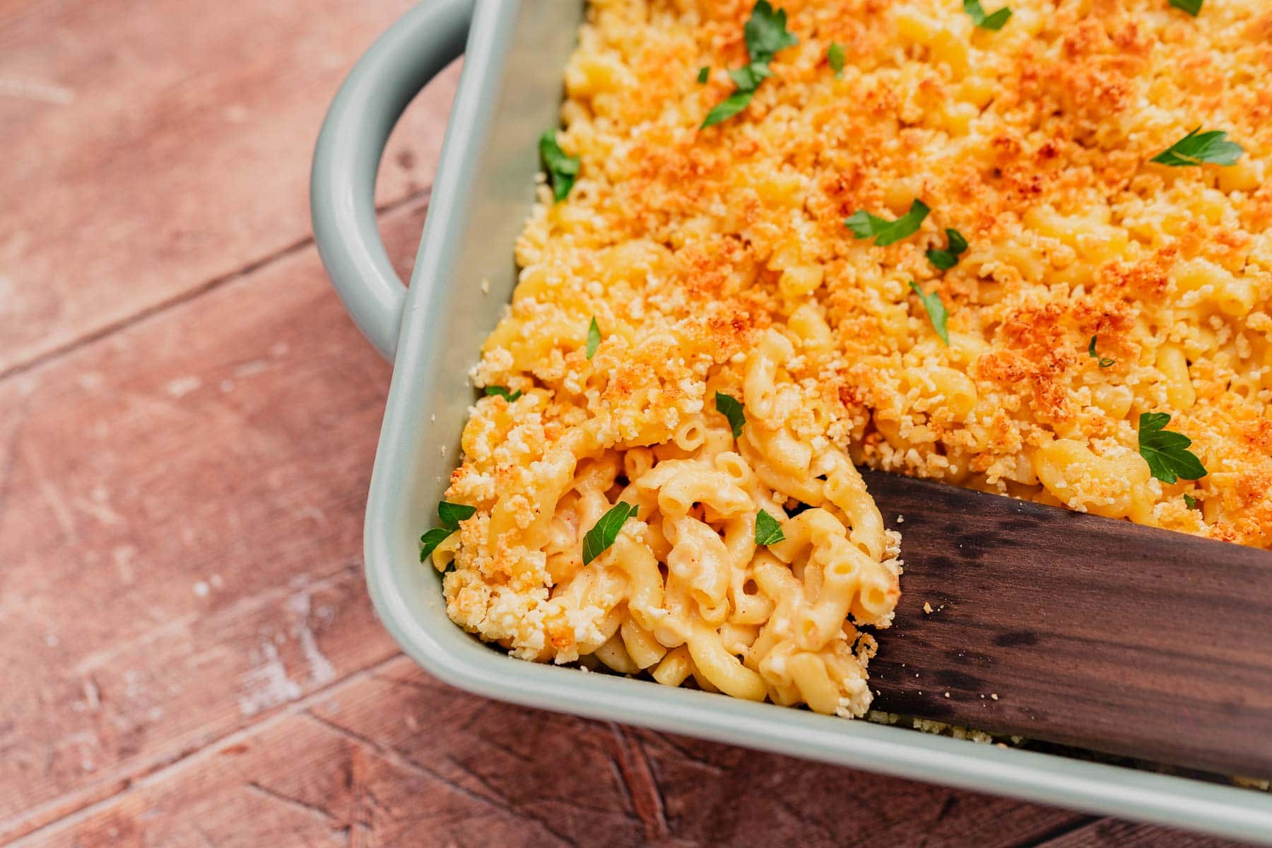 A baking dish filled with gluten free mac and cheese, topped with golden breadcrumbs and garnished with parsley, with a wooden spatula serving a portion.