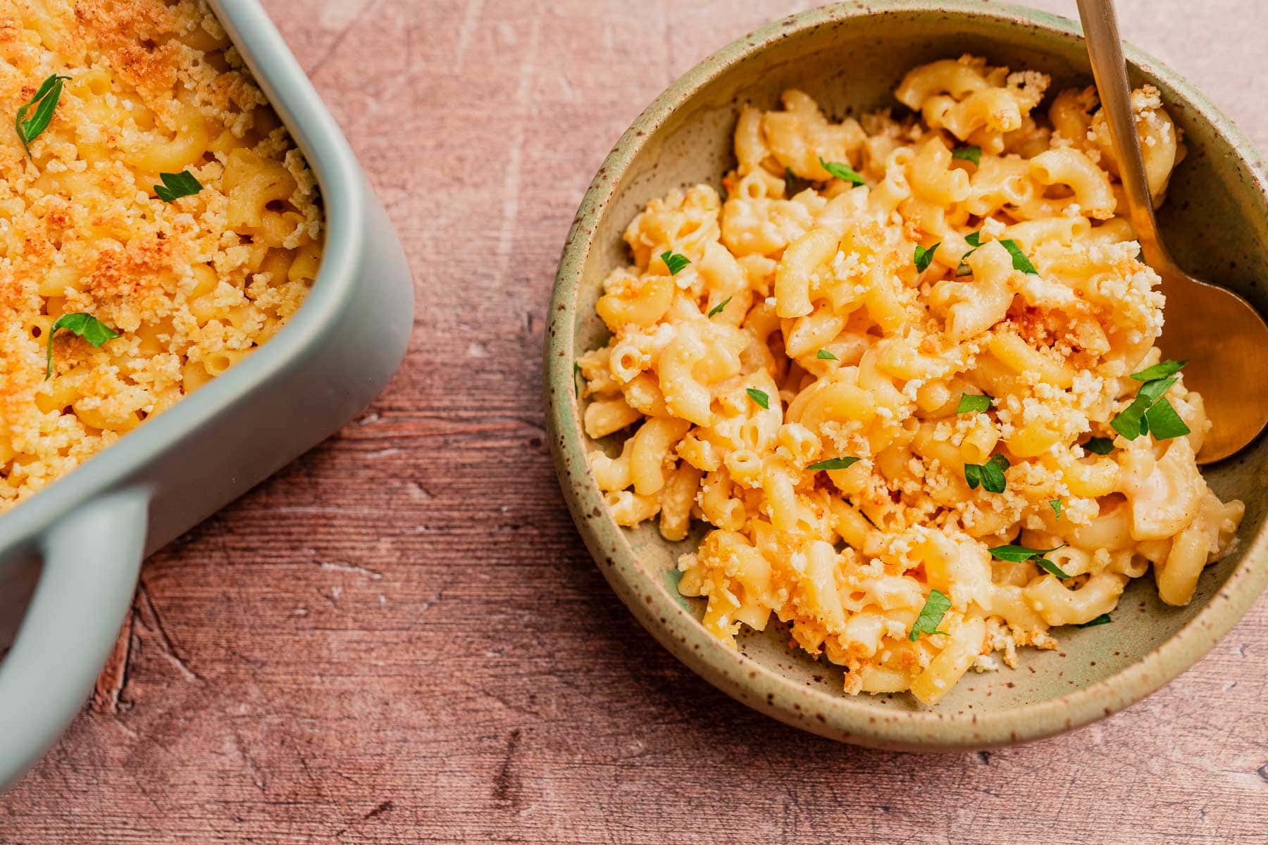 A bowl of gluten free mac and cheese topped with breadcrumbs and parsley sits next to a casserole dish of the same dish on a wooden surface.