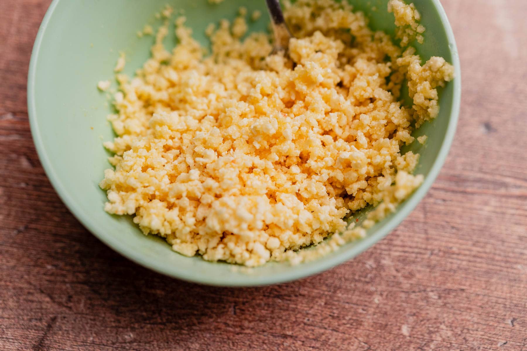 A green bowl filled with crumbled cheese or a mixture resembling ricotta or cottage cheese, perfect for making gluten free mac and cheese, sits on a wooden surface.