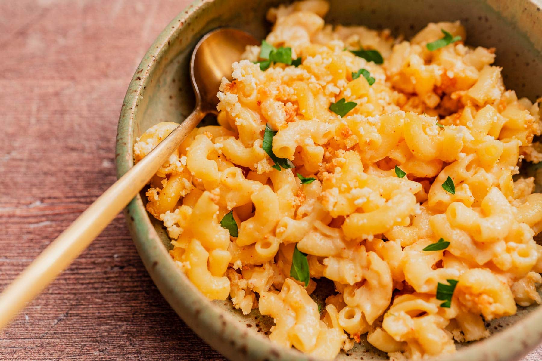 A bowl of gluten free mac and cheese topped with breadcrumbs and parsley, with a gold spoon resting in the bowl.