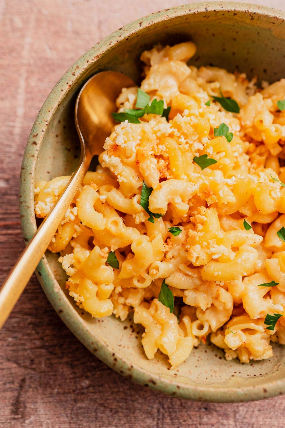 A bowl of gluten free mac and cheese topped with breadcrumbs and chopped parsley, with a gold spoon placed inside the bowl.