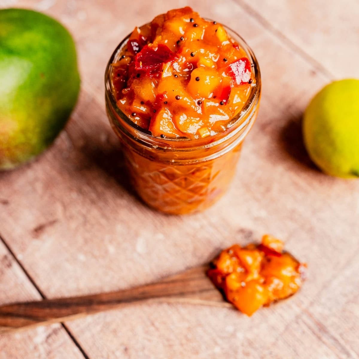 A glass jar of mango chutney sits on a wooden surface, with a wooden spoon filled with mango chutney beside it and whole mango and lime in the background.