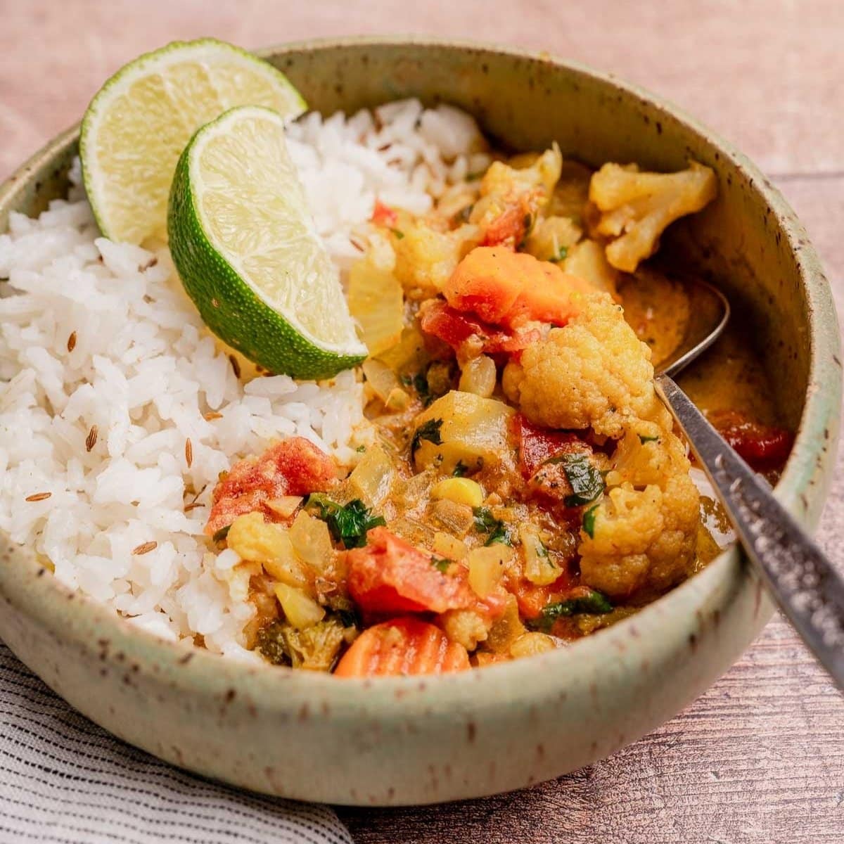 A ceramic bowl with white rice, slow cooker vegetable curry, and lime wedges on top, placed on a wooden surface with a spoon inside the bowl.