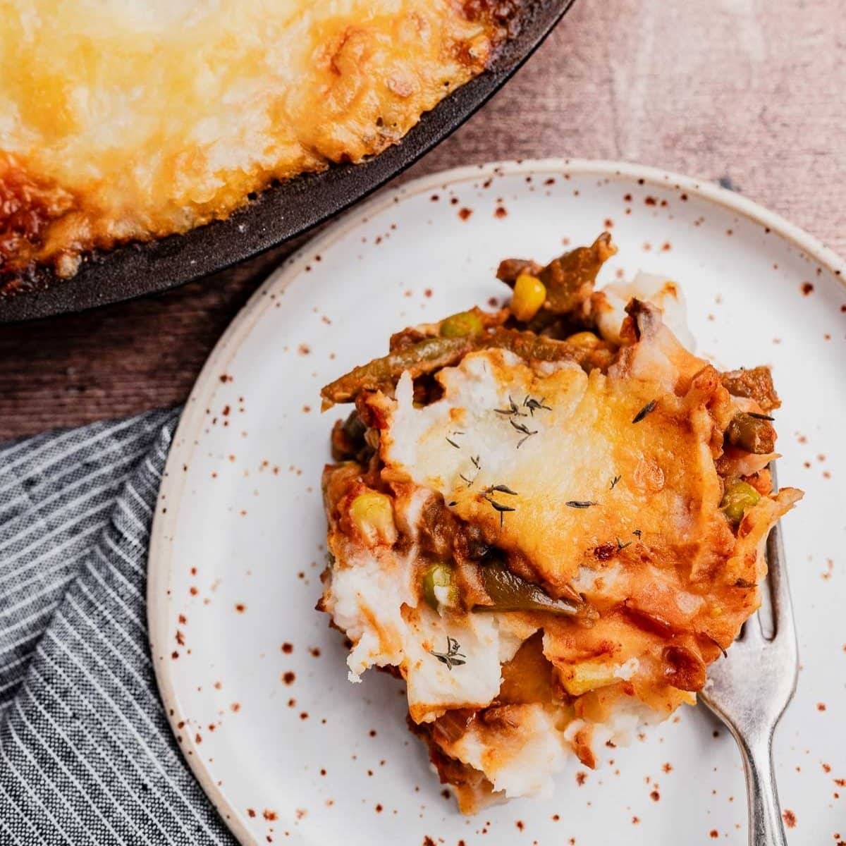 A serving of vegetarian shepherd's pie with melted cheese on top is plated with a fork beside the main dish on a wooden surface.