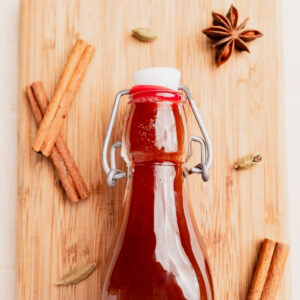 A glass bottle filled with brown chai concentrate sits on a wooden board, surrounded by cinnamon sticks, cardamom pods, and a star anise.
