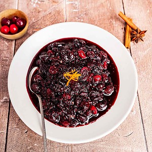 A white bowl filled with cranberry chutney garnished with orange peel, with a spoon inside. Cinnamon sticks, star anise, and a small bowl of cranberries are nearby on a wooden surface.