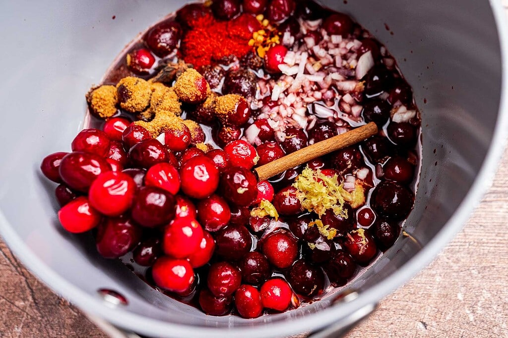 A pot filled with fresh cranberries, chopped onions, spices, ground ginger, grated lemon zest, and a cinnamon stick—perfect ingredients for a flavorful cranberry chutney, ready to be mixed and cooked.