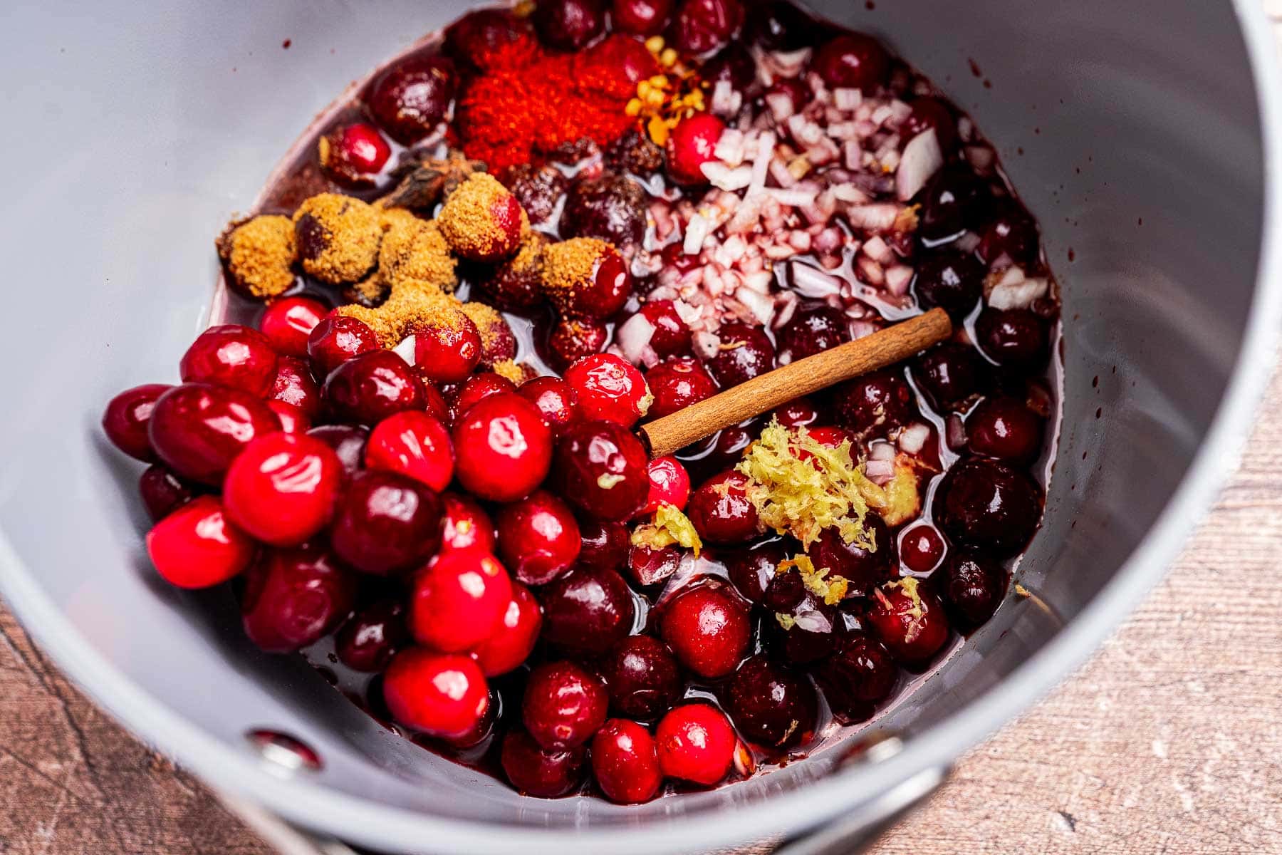 A pot filled with fresh cranberries, chopped onions, spices, ground ginger, grated lemon zest, and a cinnamon stick—perfect ingredients for a flavorful cranberry chutney, ready to be mixed and cooked.