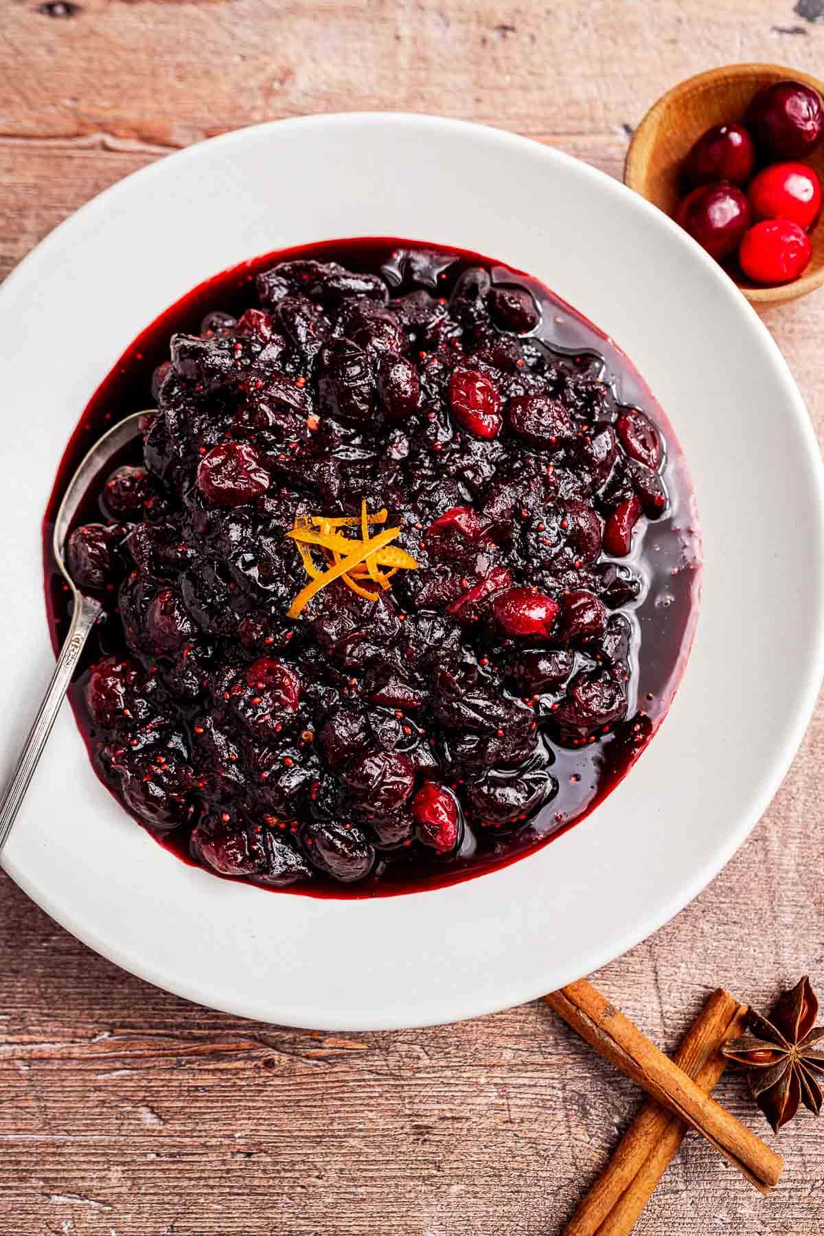A white bowl filled with cranberry chutney sauce garnished with orange zest, with a spoon, whole cranberries, and cinnamon sticks beside it on a wooden surface.