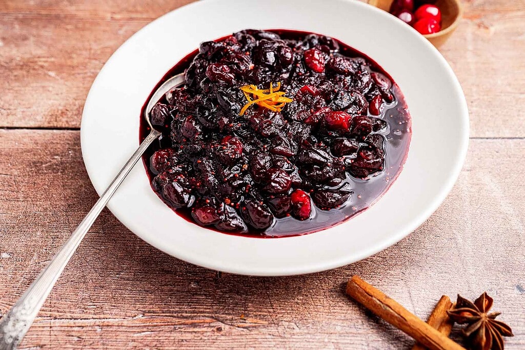 A white plate with homemade cranberry chutney, garnished with orange zest, sits beside a cinnamon stick and star anise on a wooden table.
