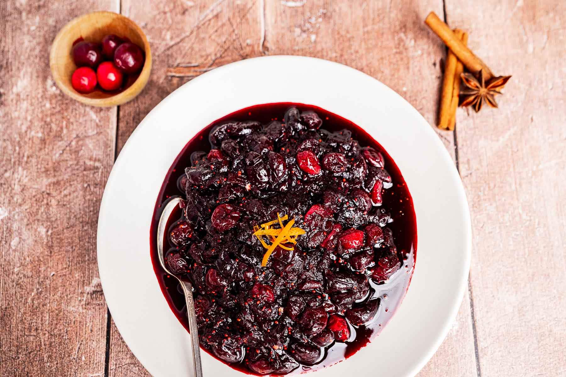 A bowl of cranberry chutney with orange zest on top, a spoon in the bowl, and a dish of cranberries, cinnamon sticks, and star anise beside it on a wooden surface.