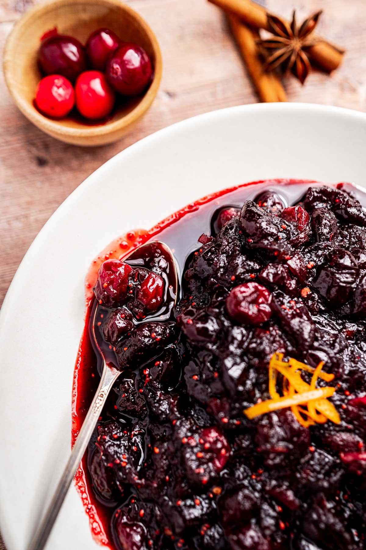 A bowl of cranberry chutney garnished with orange zest, with a spoon, and a small wooden bowl of fresh cranberries in the background.