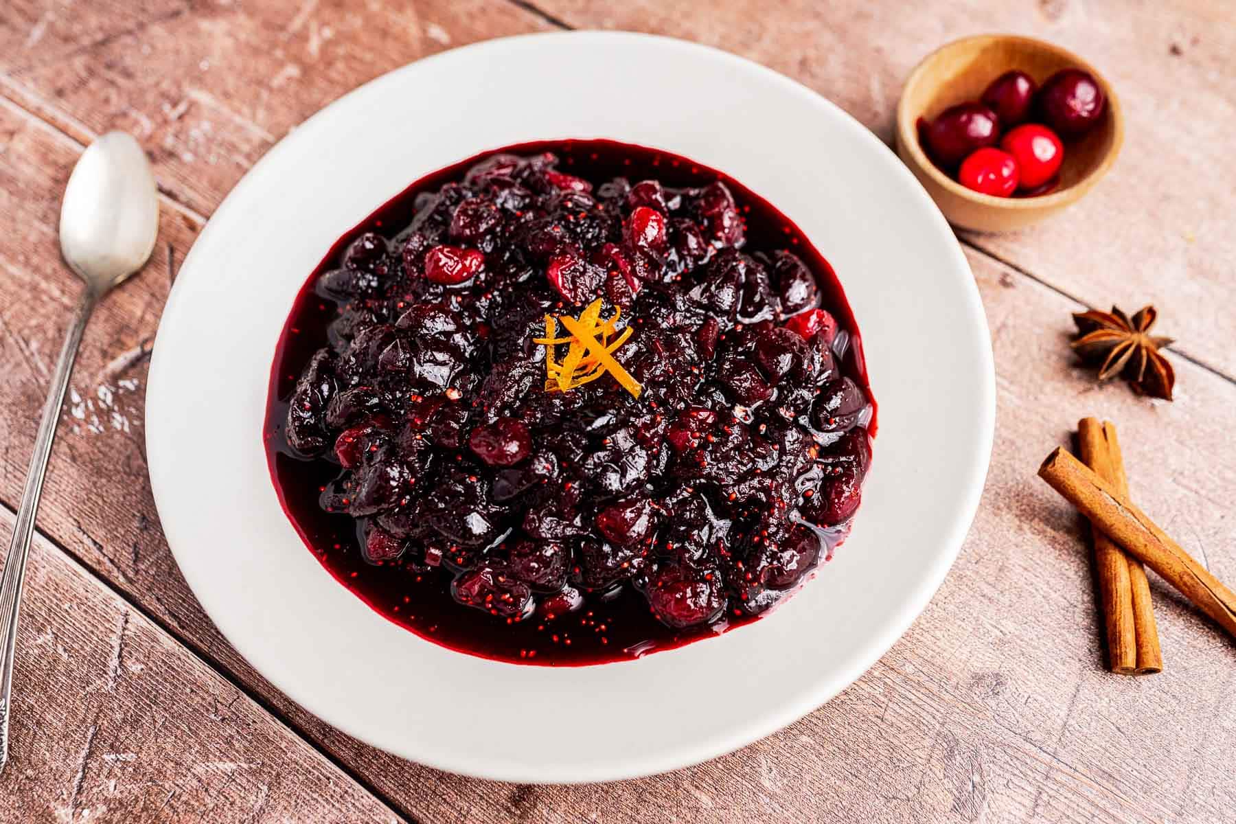 A white plate of cranberry chutney garnished with orange zest, with a spoon, cinnamon sticks, a star anise, and a small bowl of cranberries on a wooden table.