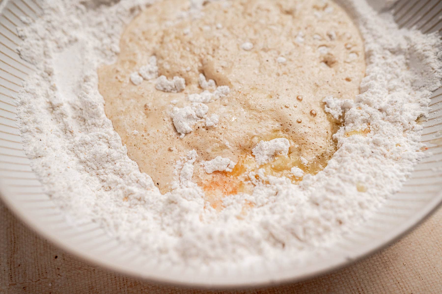 A close-up of gluten free dinner rolls ingredients—flour, yeast mixture, and an egg—in a white mixing bowl, prepared for baking.