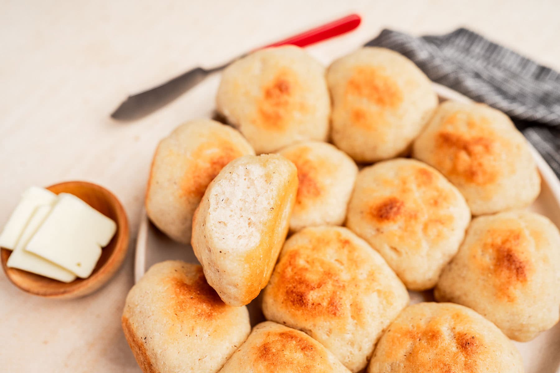 A plate of gluten free dinner rolls with a single roll split open, next to a small bowl of butter slices and a butter knife on a napkin.