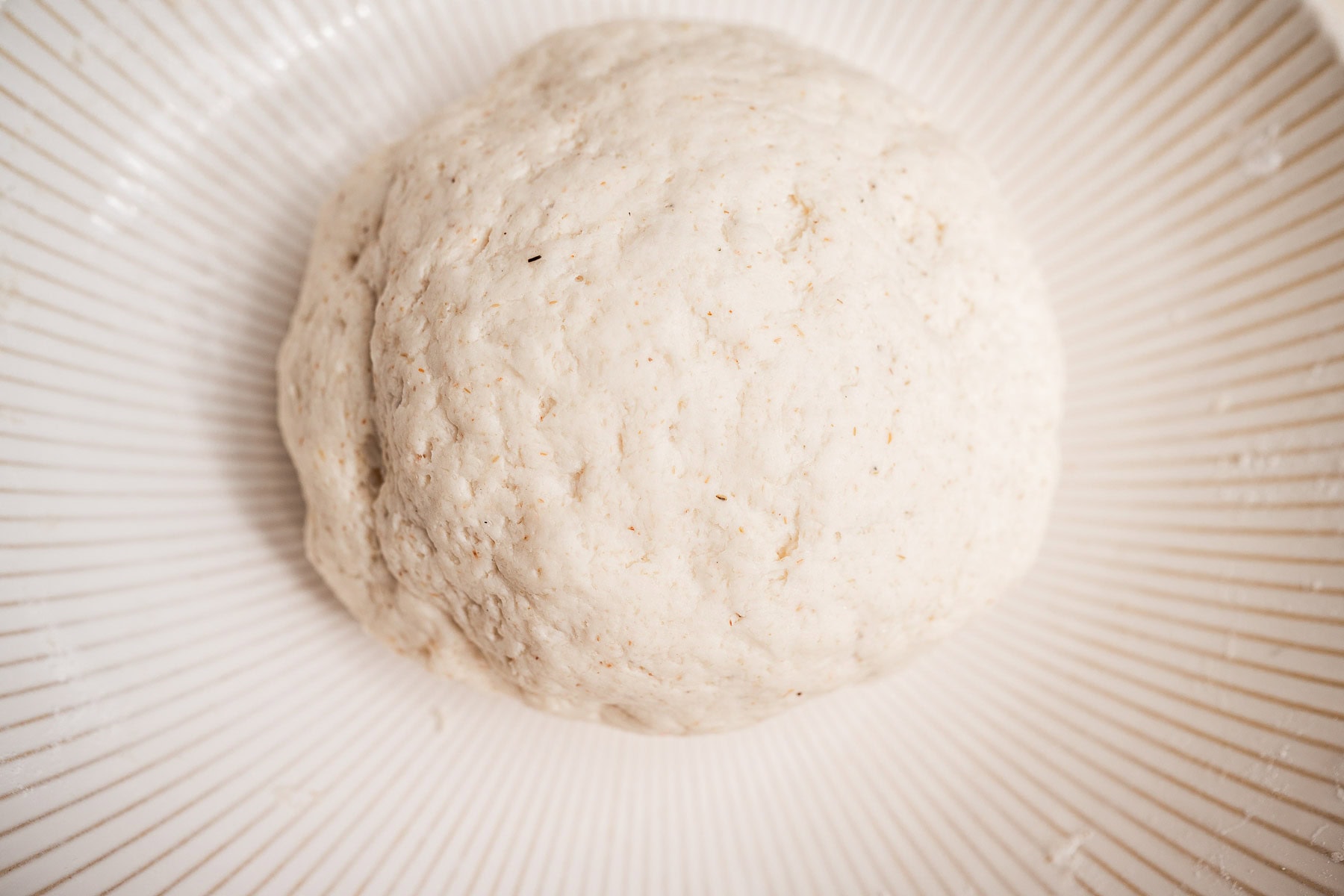 A ball of raw dough sits on a ribbed white plate, ready for preparation or baking into soft, gluten free dinner rolls.