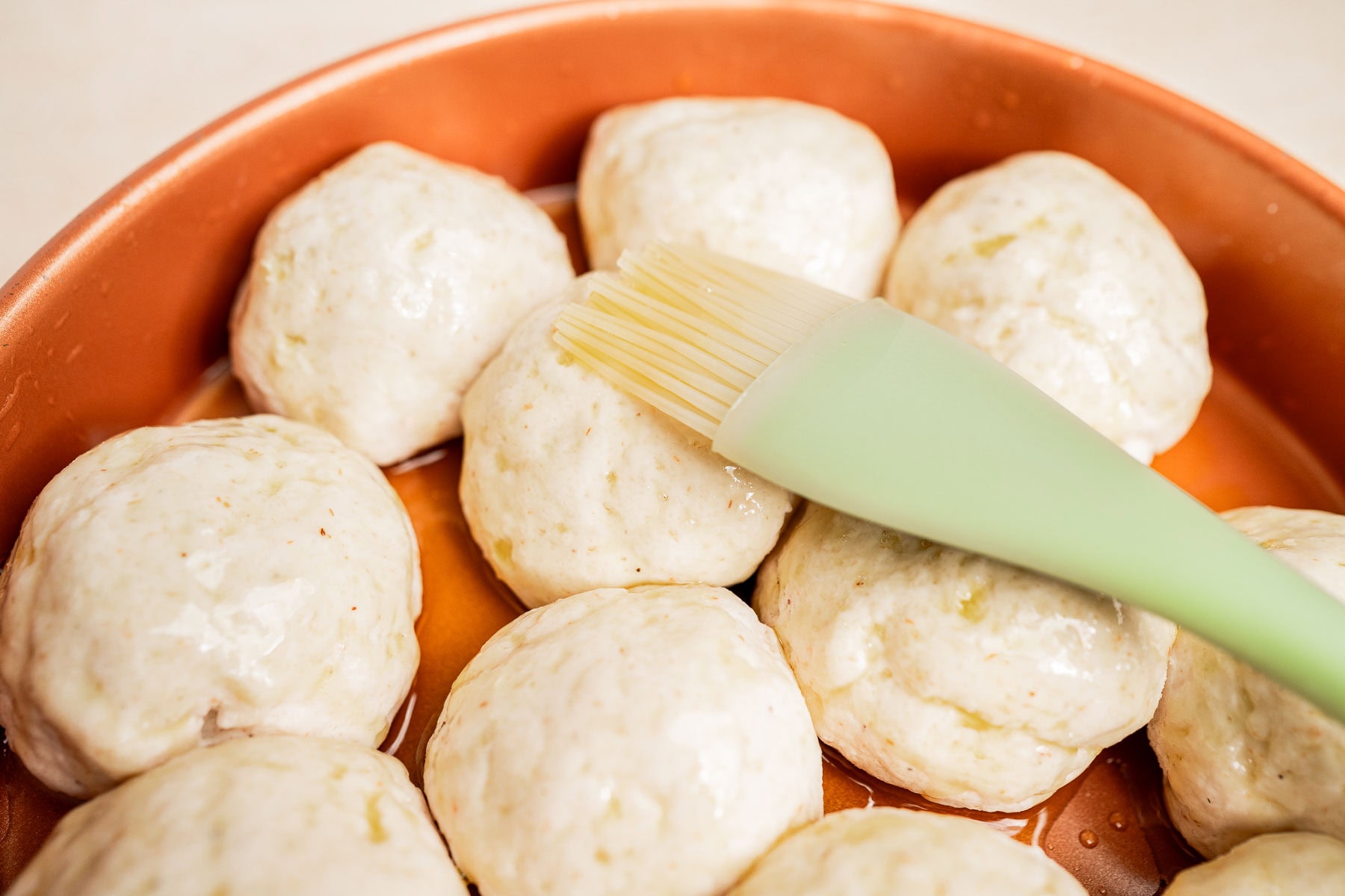 A green silicone brush is being used to coat raw gluten free dinner rolls in oil or butter on a round copper-colored baking tray.
