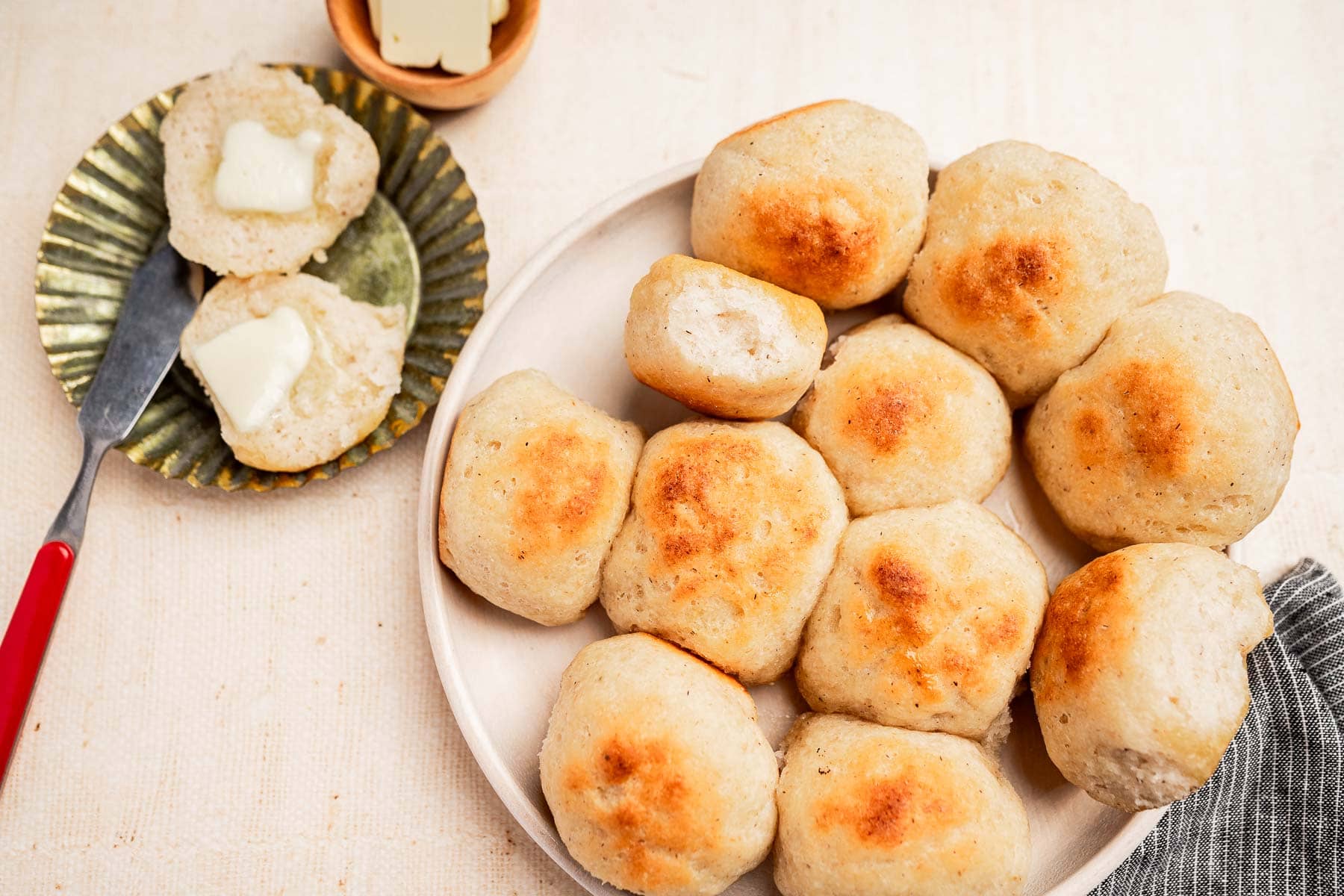 A plate of golden brown gluten free dinner rolls with one split open, served alongside a plate with two buttered halves and a butter knife.