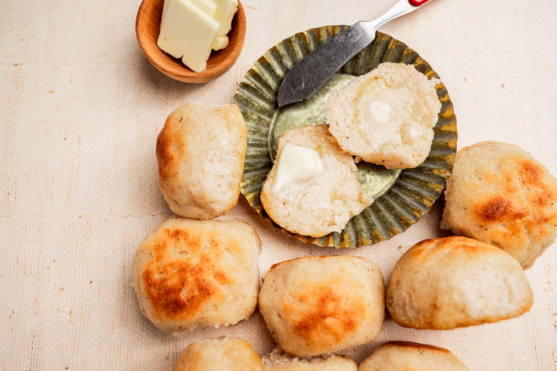 A plate with homemade gluten free dinner rolls, one split open with a pat of melting butter. A butter knife and a small bowl of butter are beside the rolls.