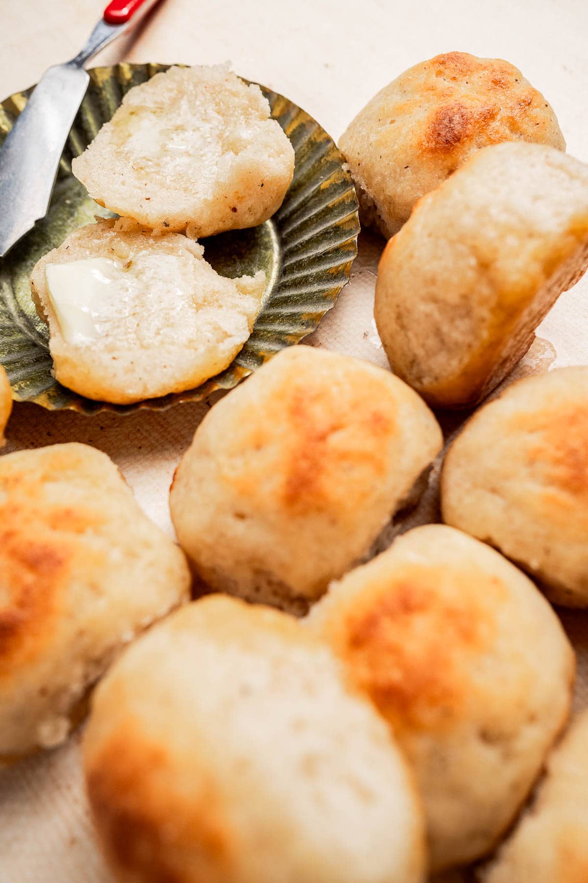 Close-up of several golden brown gluten free dinner rolls, with one roll split open and spread with butter on a plate beside a butter knife.