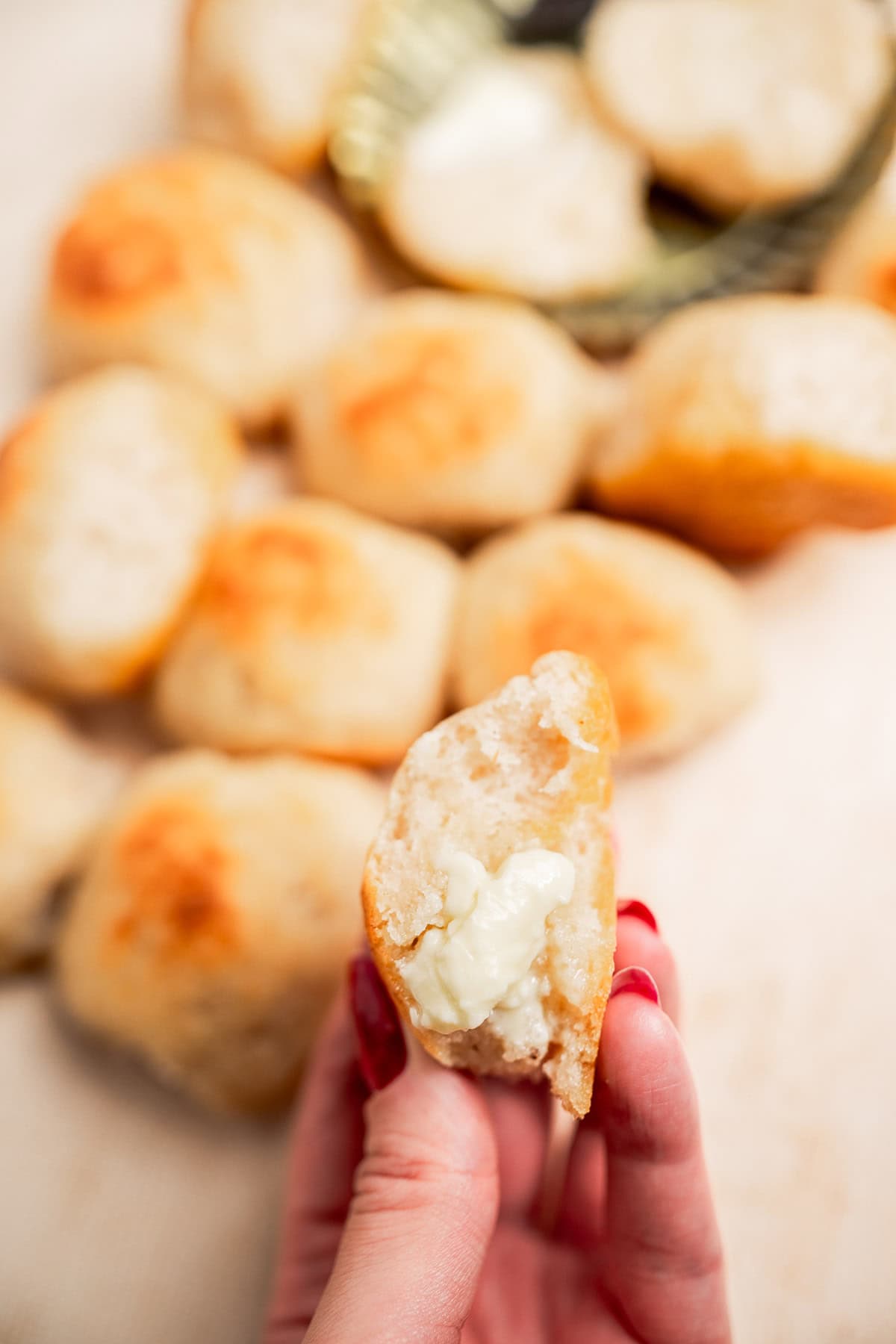 A hand holds a piece of gluten free dinner roll with melted butter inside; more bread rolls and butter are visible in the background.