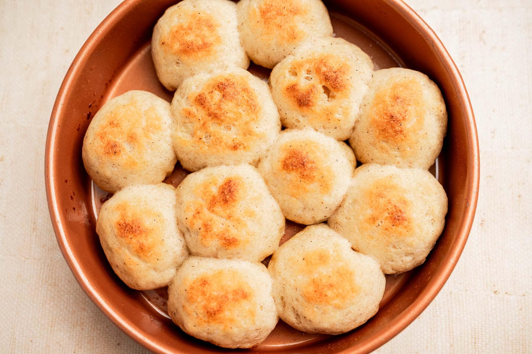Round, golden-brown gluten free dinner rolls are arranged closely together in a circular brown baking dish.