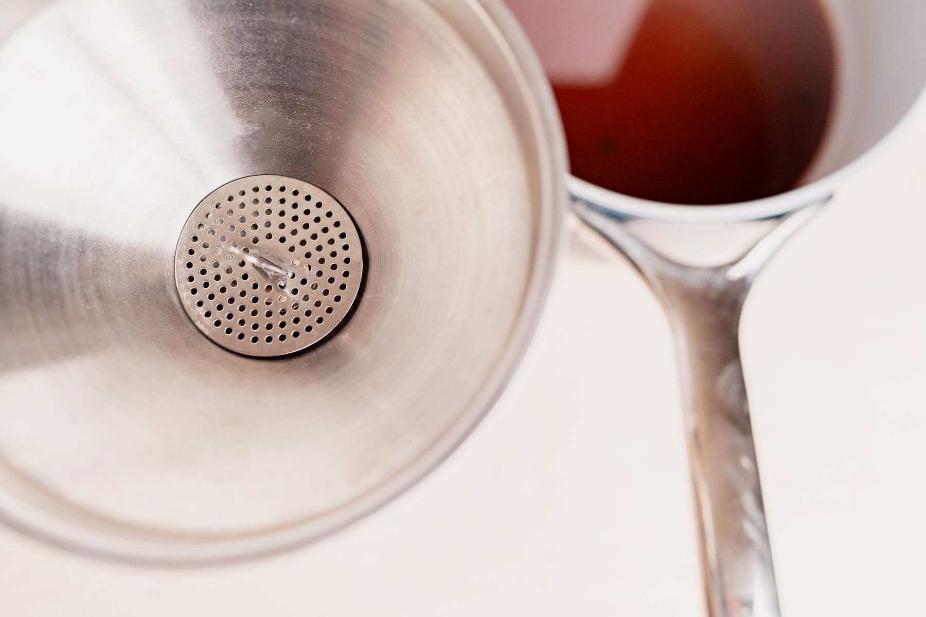 A metal funnel with a strainer sits next to a saucepan filled with chai concentrate, its rich brown liquid seen from above.
